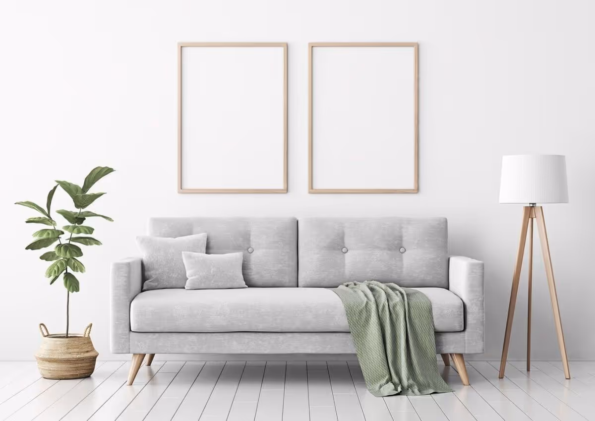 Minimalist living room with a light gray sofa, two blank wooden frames above, a potted plant on the left and a tripod floor lamp on the right.