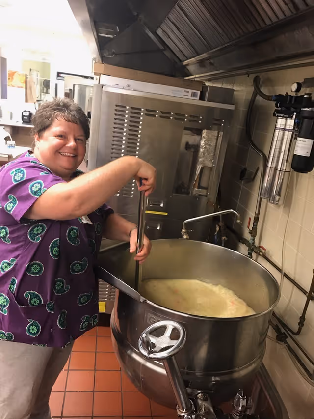 A person smiling while stirring a large industrial kettle of soup in a commercial kitchen.