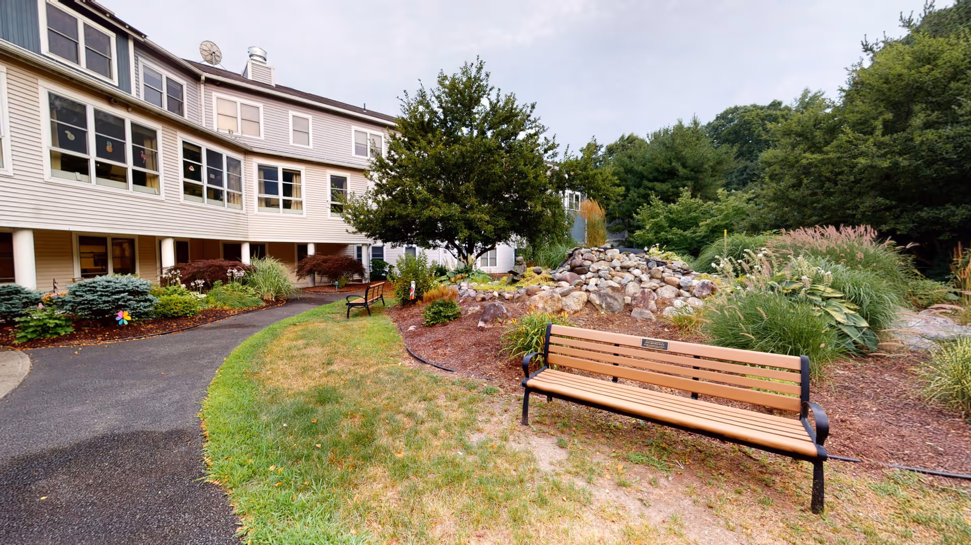 Outdoor garden area at Ruth's House featuring a paved walkway, green grass, two wooden benches, a small rock formation, various plants and trees, and a multi-story building with large windows in the background.