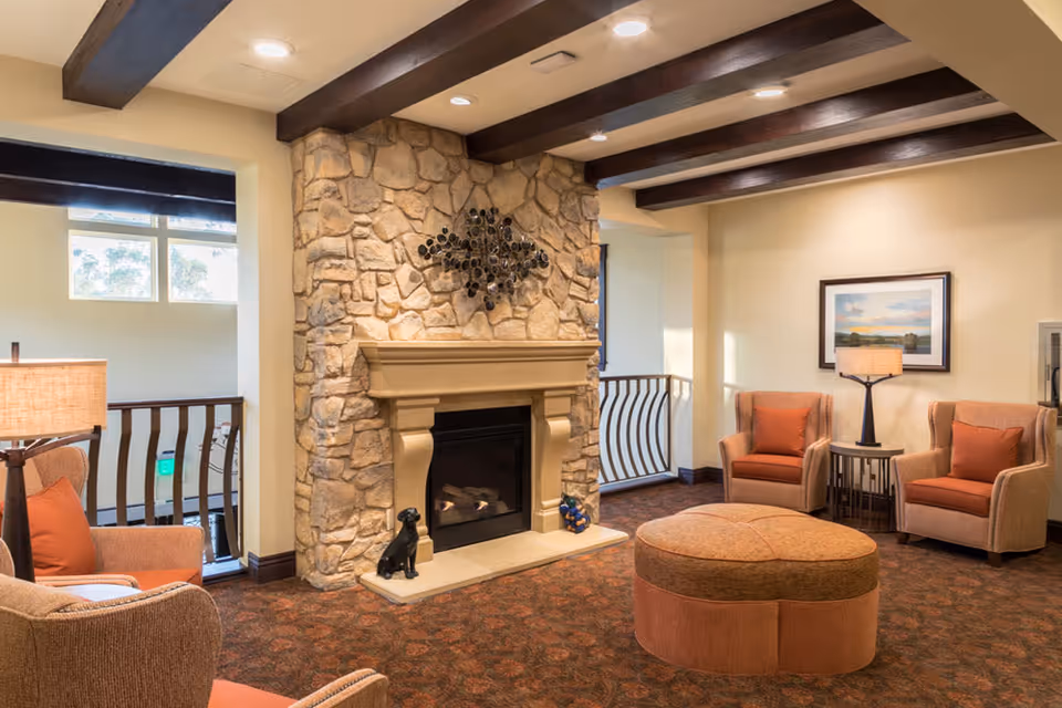 A cozy living room area with a stone fireplace in the center, decorated with a metal wall art piece above the mantel. The room features four cushioned armchairs with orange pillows arranged around a large, round ottoman. There are two table lamps on side tables, a framed landscape painting on the wall, and exposed wooden beams on the ceiling. A railing and windows are visible in the background.