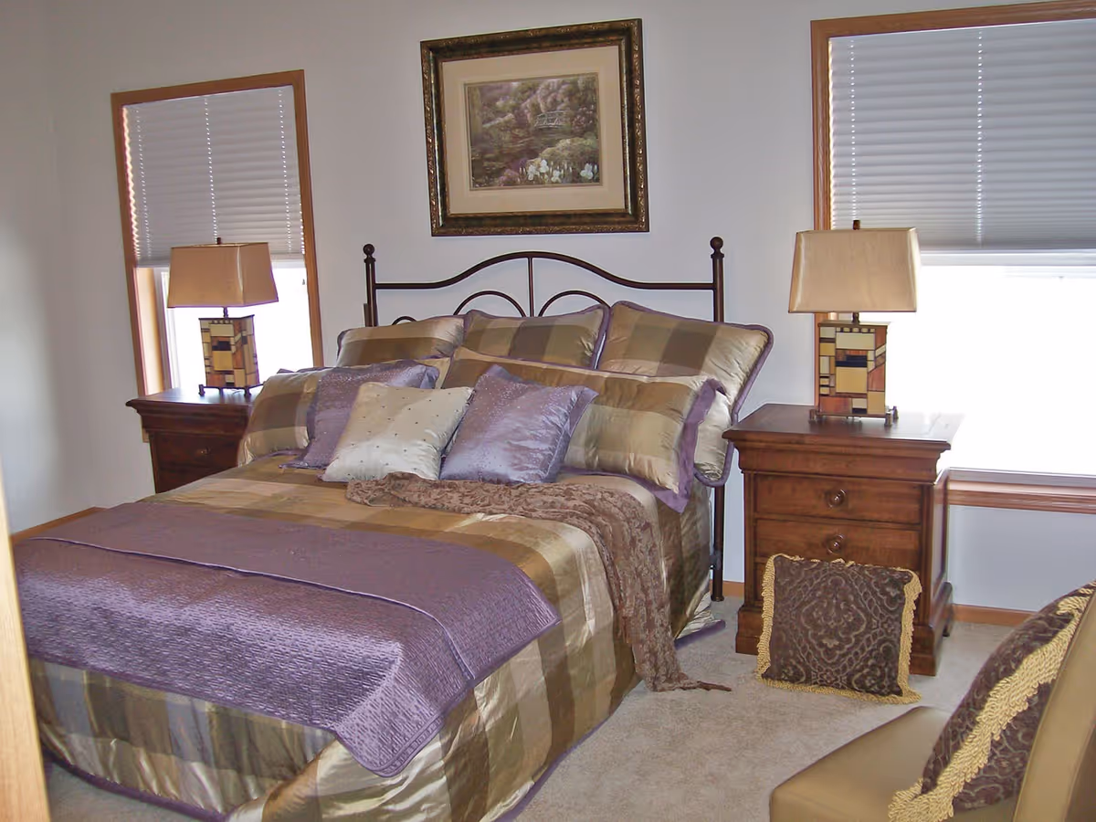 A neatly made bed with a metal headboard in a bedroom. The bed has a checkered bedspread in shades of brown and purple, with multiple pillows in coordinating colors. On each side of the bed are wooden nightstands with matching table lamps. There are two windows with blinds partially closed, and a framed picture hanging on the wall above the bed.