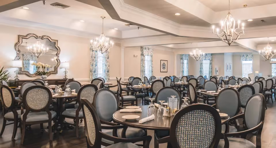 A spacious dining room with multiple round tables set with plates, napkins, and glasses. The room features elegant chandeliers, large decorative mirrors, floral curtains on the windows, and upholstered chairs with wooden frames.