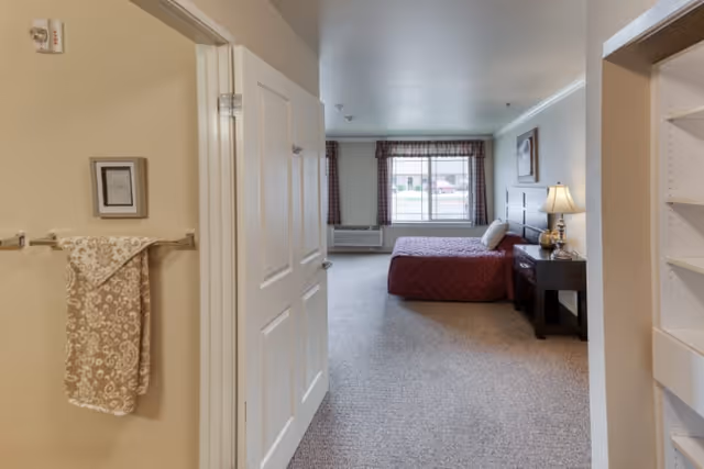 View into a senior living bedroom from an adjacent hallway. The bedroom features a bed with a red quilt, a nightstand with a lamp, a window with plaid curtains, and a framed picture on the wall. The hallway has a towel hanging on a rack and built-in shelving on the right side.