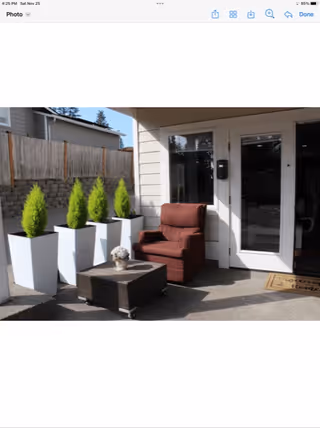 Outdoor patio area with a brown cushioned armchair, a small square coffee table on wheels with a decorative item on top, four tall white planters with green plants, a glass door entrance, and a welcome mat in front of the door.