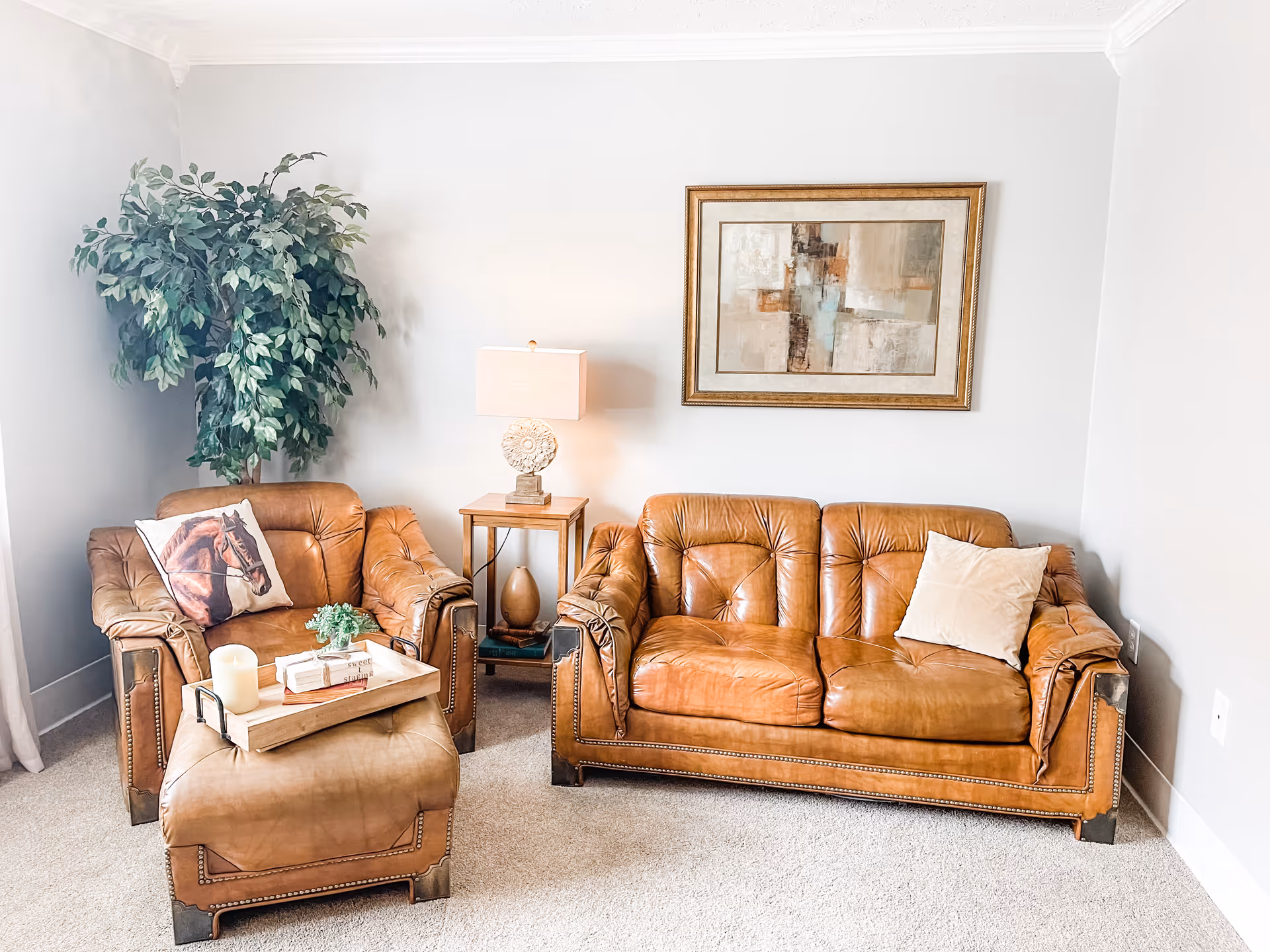 Cozy living room with tan leather sofa and armchair, an ottoman with a tray, side table with a lamp, a potted plant, and framed artwork on a light gray wall.