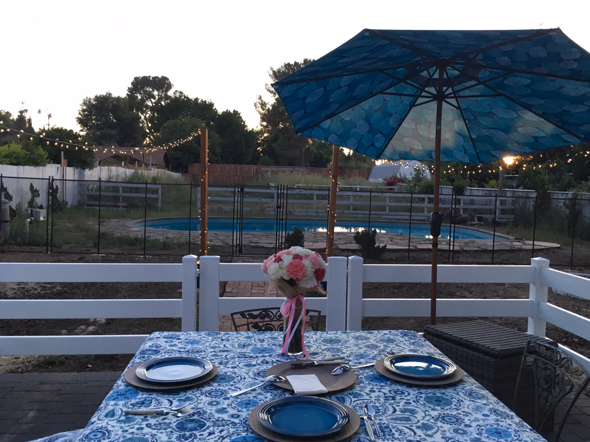 Outdoor patio dining table with blue patterned tablecloth, place settings and a floral centerpiece under a blue umbrella, overlooking a fenced backyard pool.