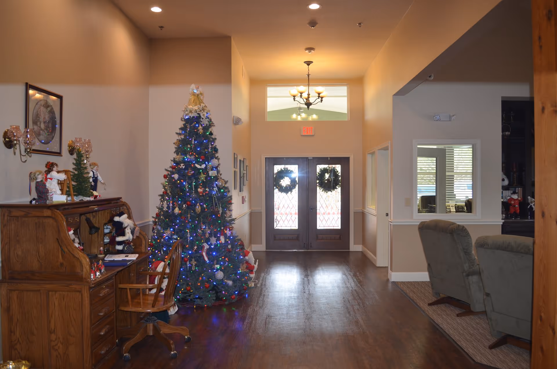 Interior view of a senior living facility hallway decorated for Christmas with a large Christmas tree adorned with lights and ornaments. To the left, there is a wooden desk with dolls and small Christmas decorations. At the end of the hallway, double doors with glass panels have wreaths hanging on them. To the right, there are two green upholstered chairs and a window opening into another room.