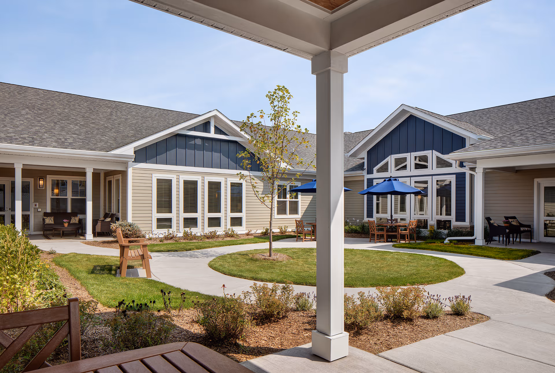 Outdoor courtyard area at Heritage Woods of McHenry featuring a circular concrete walkway surrounding a small grassy area with a young tree in the center. The courtyard is bordered by single-story buildings with beige and blue siding, multiple windows, and covered patios with seating and tables shaded by blue umbrellas. The sky is clear and blue.
