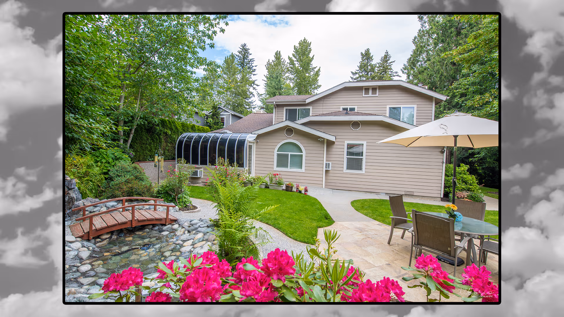 A serene outdoor garden area at Kenmore Retreat Adult Family Home featuring a small wooden bridge over a rocky stream, vibrant pink flowers in the foreground, a patio with a table, chairs, and a large umbrella, surrounded by lush green trees and shrubs, with a beige two-story building in the background.