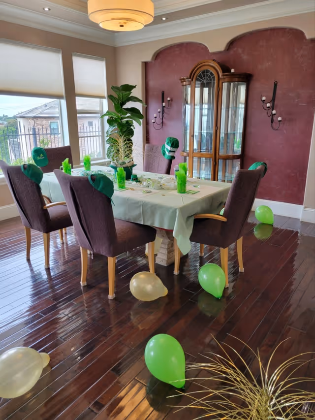 Dining room with a decorated table, green balloons on the floor, purple chairs, and a glass-front china cabinet against a maroon wall.