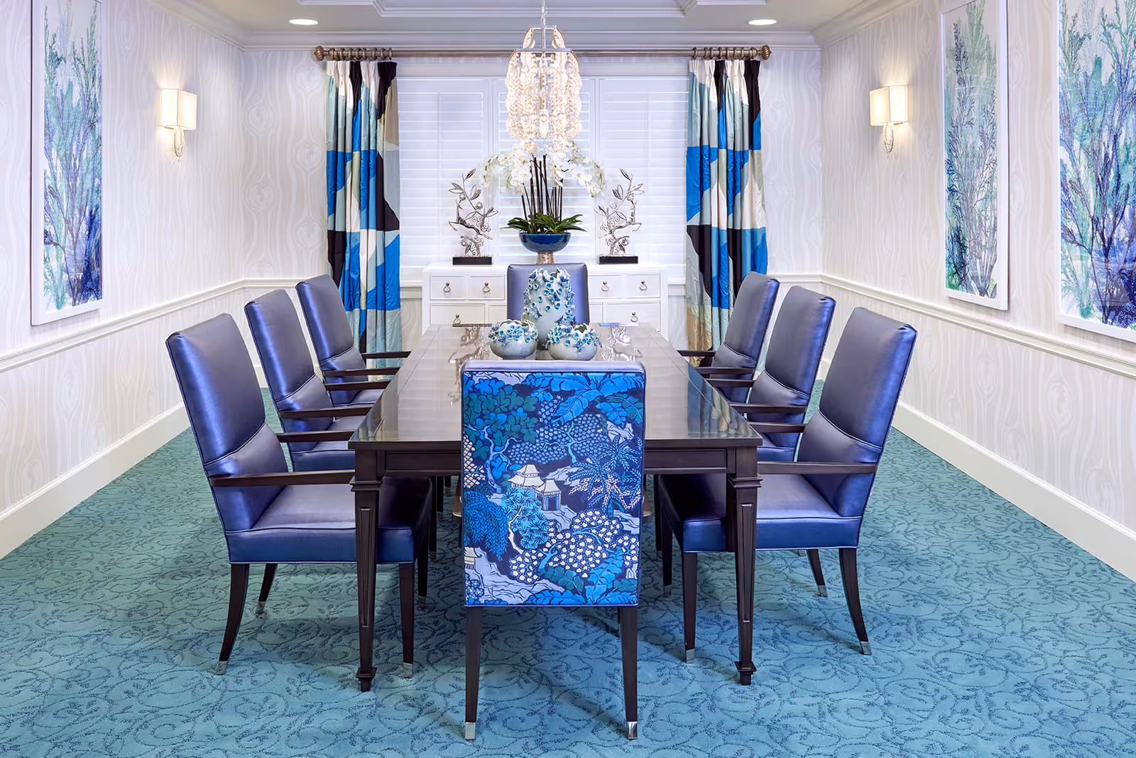 A formal dining room with a long dark wooden table surrounded by eight blue upholstered chairs, one of which has a patterned blue backrest. The room features light patterned wallpaper, teal carpet with a subtle design, two large colorful paintings on the walls, blue and white curtains, a white sideboard with decorative items, and a chandelier hanging above the table.