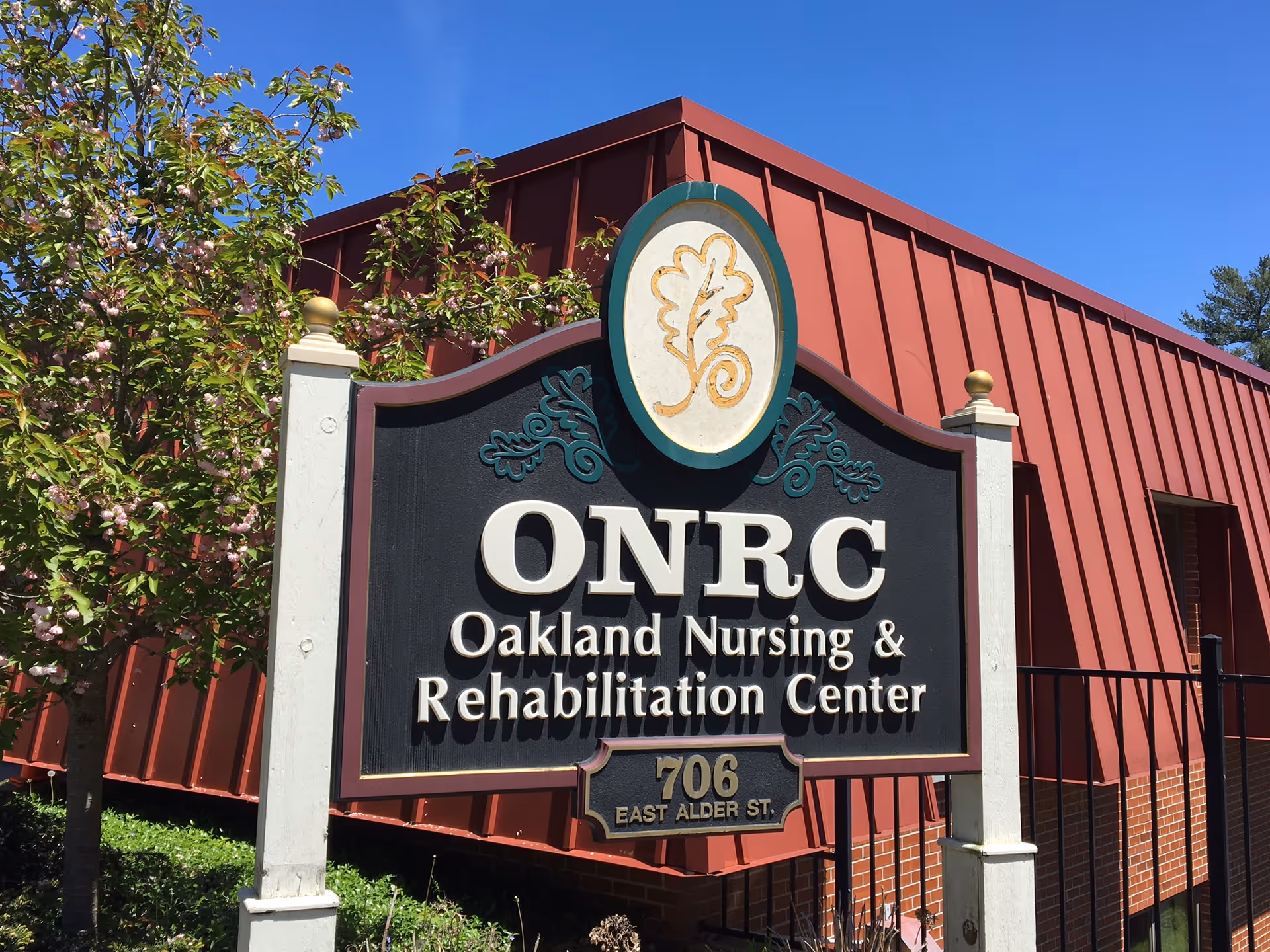 A sign for Oakland Nursing & Rehabilitation Center with the abbreviation ONRC, located at 706 East Alder St. The sign is mounted on white posts with a decorative emblem at the top, and a red building with a slanted roof is visible in the background along with a tree with green leaves and pink flowers under a clear blue sky.