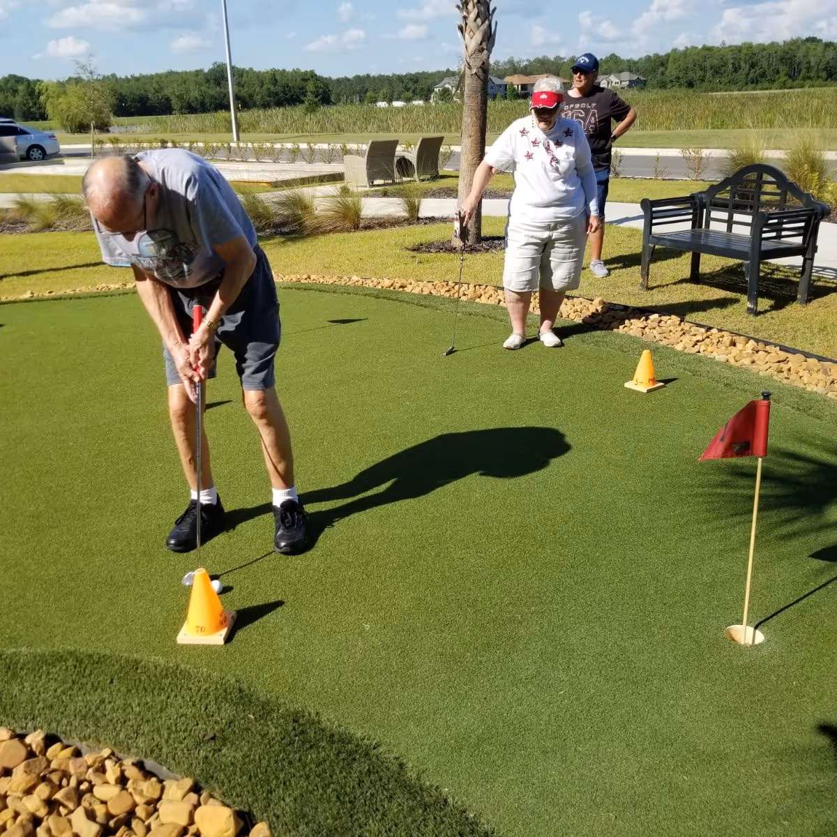 Three elderly people playing mini golf on a putting green outdoors on a sunny day. One man is putting the ball while a woman and another man watch. There is a bench, palm tree, and some chairs in the background with a grassy area and a road beyond.