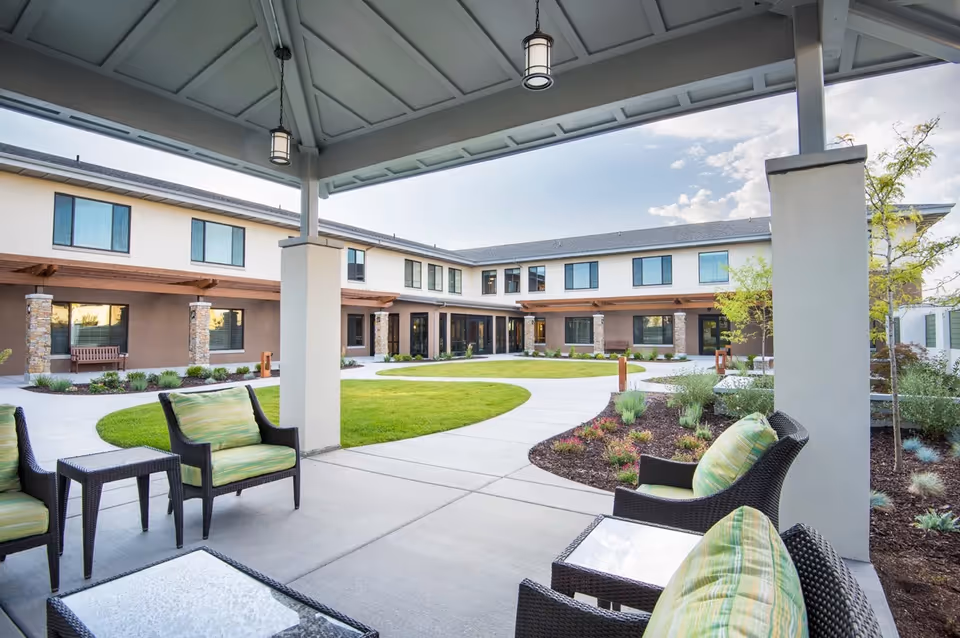Covered patio with wicker chairs and green cushions overlooking a landscaped courtyard and a two-story senior living building.