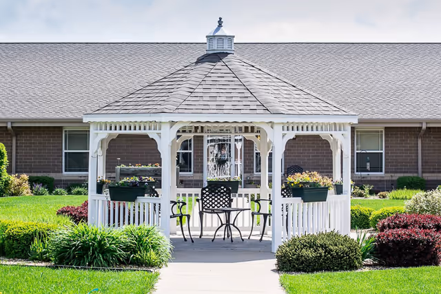 A white wooden gazebo with a table and chairs in a landscaped courtyard in front of a single-story brick building.