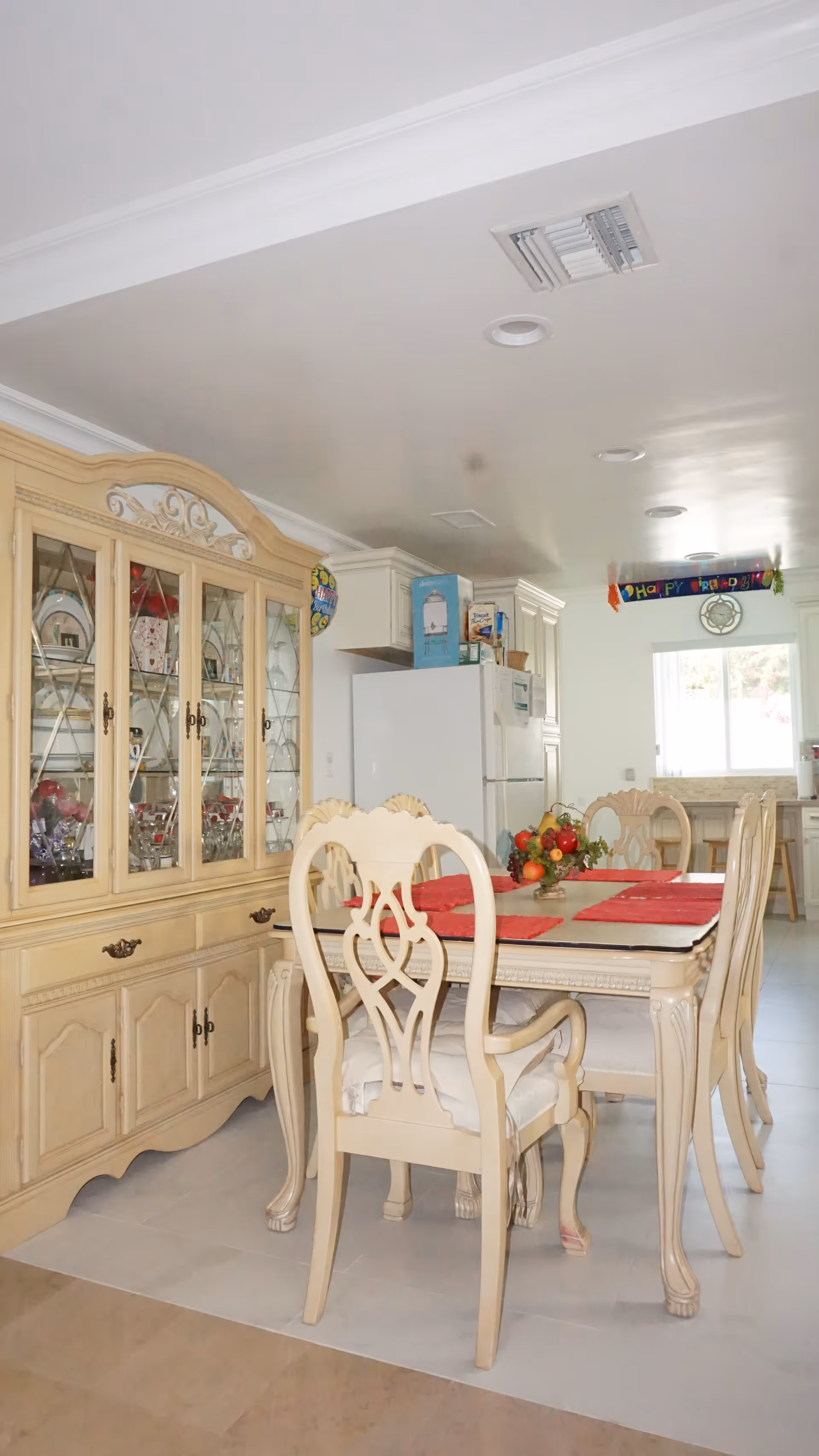 A dining area with a cream-colored wooden dining table and six matching chairs with cushioned seats. The table is set with red placemats and a fruit centerpiece. Behind the table is a large matching china cabinet filled with glassware and dishes. In the background, there is a white refrigerator and kitchen cabinets. A 'Happy Birthday' banner is hanging near the ceiling, and a window lets in natural light.