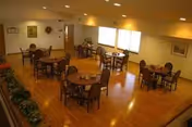Senior living dining room with round tables, chairs, warm wooden floor, soft lighting.
