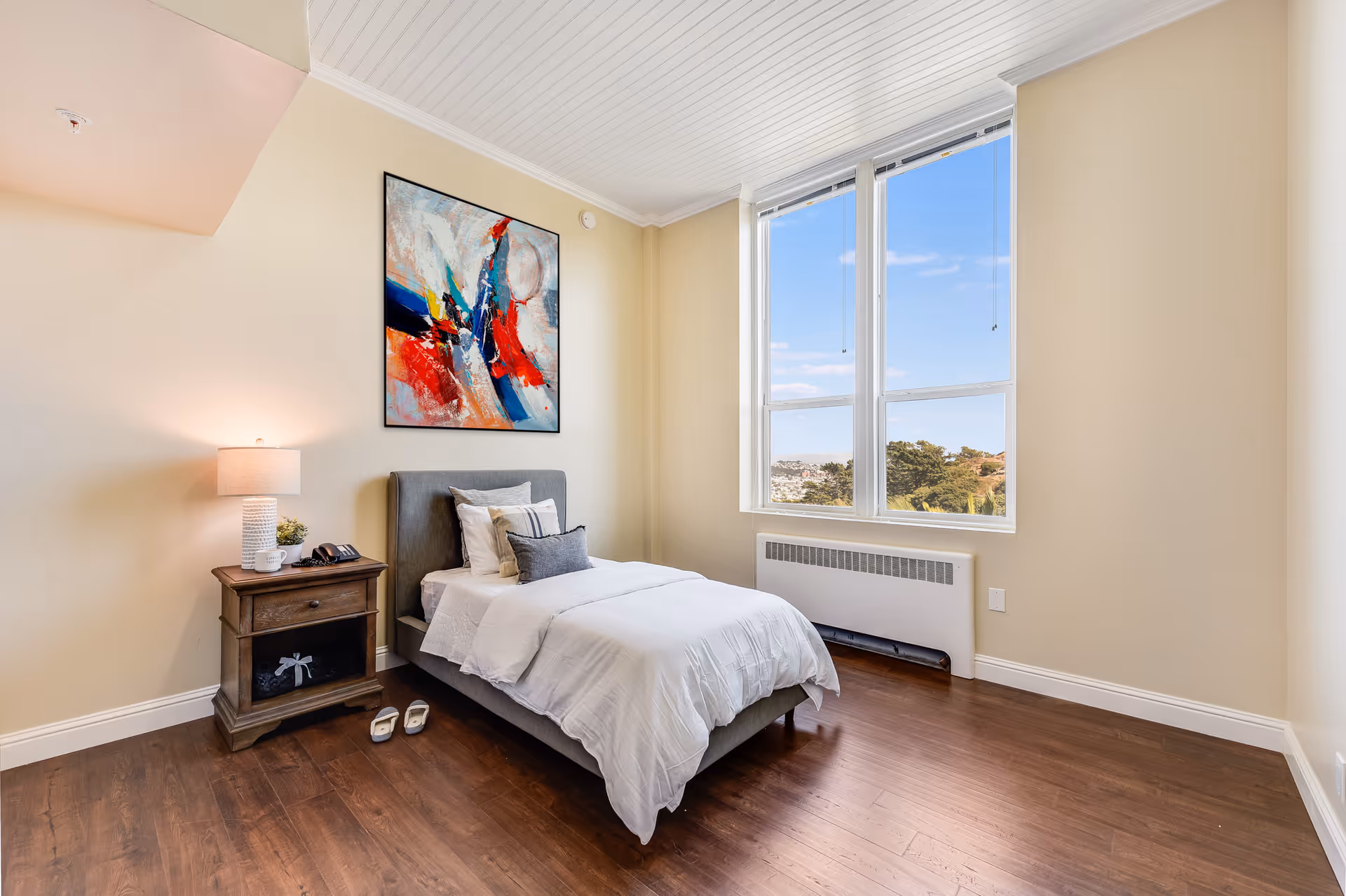 A bright bedroom with a single bed made with white bedding and multiple pillows. Next to the bed is a wooden nightstand with a lamp, a small plant, a phone, and a coffee mug. Above the bed hangs a colorful abstract painting. The room has large windows showing a view of trees and a blue sky outside. The floor is wooden, and the walls are painted light beige.
