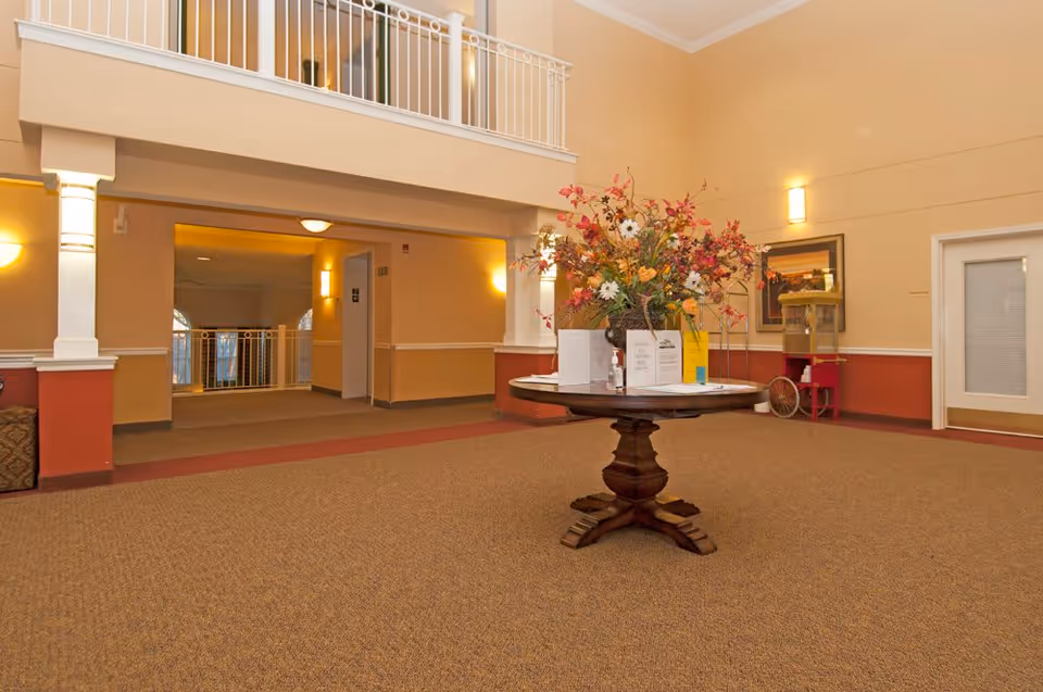 Spacious senior living lobby with a central round table holding a large floral arrangement, warm lighting, and an upstairs railing.