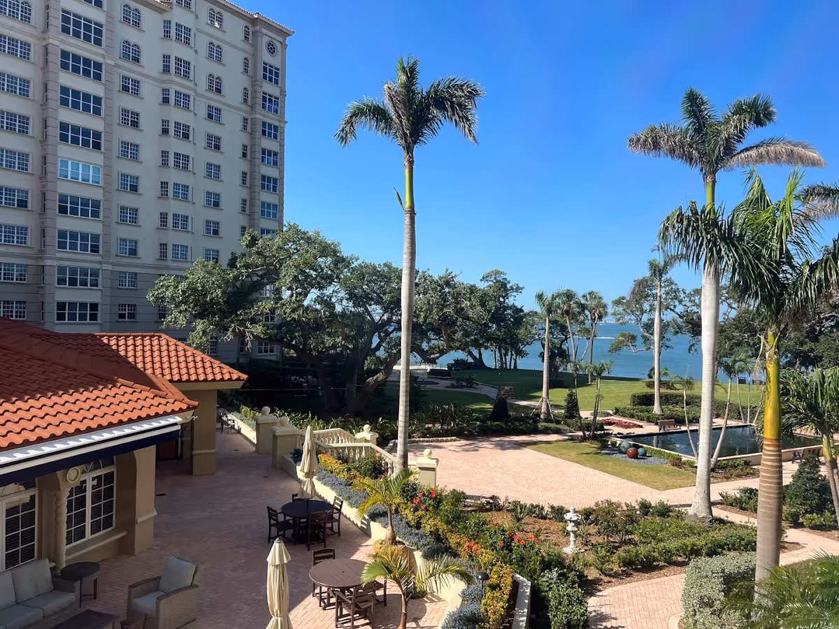 Outdoor view of The Inn at Sarasota Bay Club featuring a multi-story building with numerous windows, a courtyard with tables, chairs, umbrellas, palm trees, landscaped gardens, and a water body in the background under a clear blue sky.