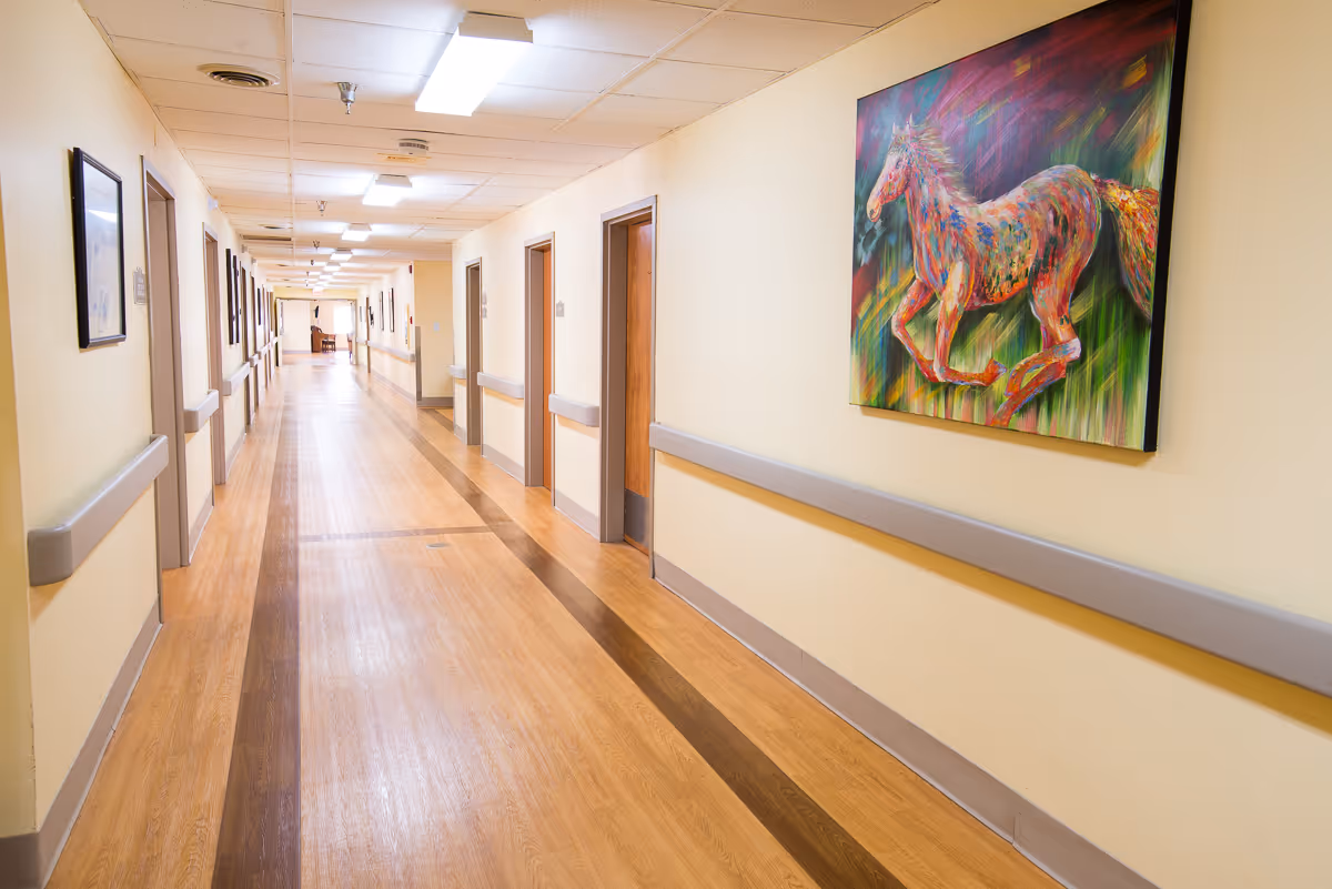 Long, well-lit interior hallway of a rehab facility with wood-look floors, handrails, multiple doors, and a colorful horse painting on the wall.