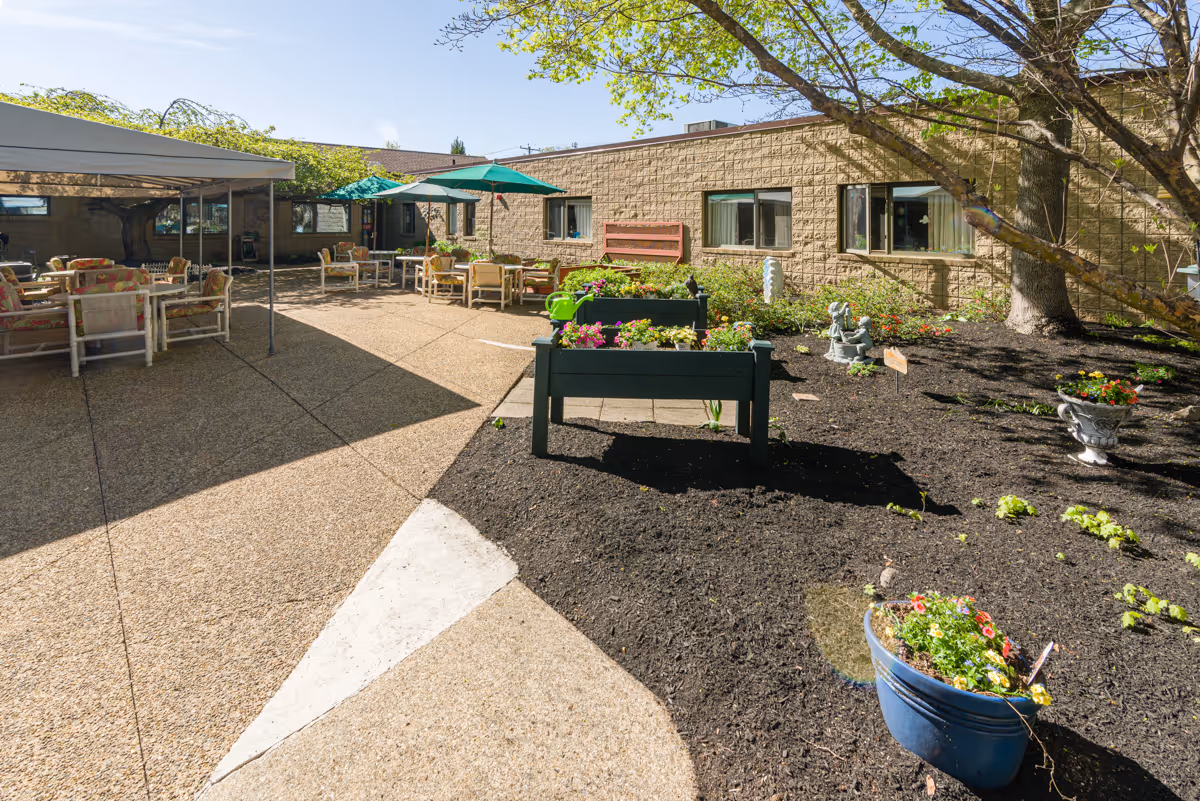 Outdoor courtyard area with garden beds filled with flowers and plants, patio tables with umbrellas and chairs, and a shaded seating area. The courtyard is surrounded by a single-story building with windows and trees providing additional shade.