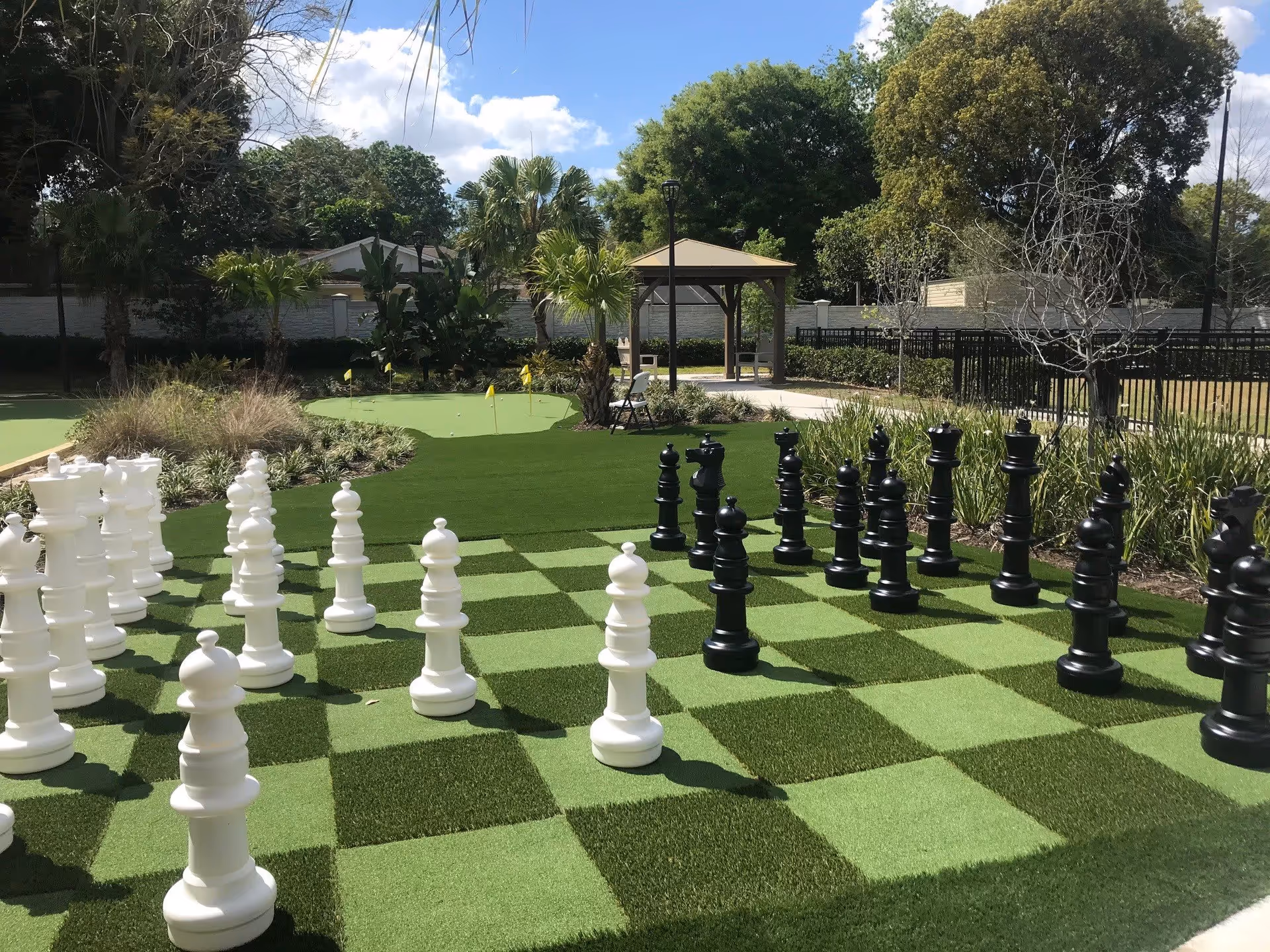 Outdoor area with a large chessboard featuring oversized black and white chess pieces on a green checkered surface. In the background, there is a putting green with small yellow flags, surrounded by plants, trees, and a gazebo under a partly cloudy sky.