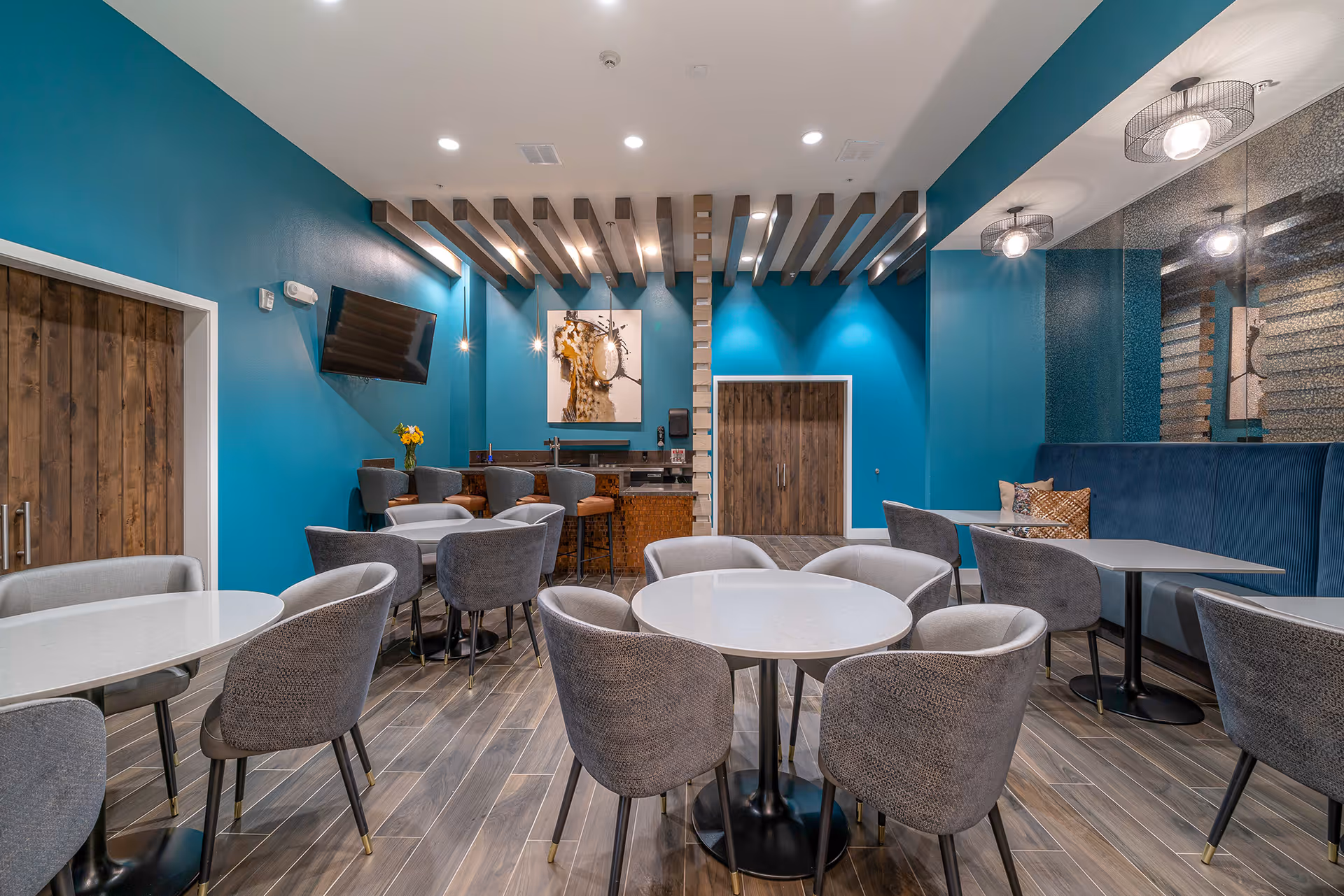 Interior view of a modern dining area with teal walls, round and rectangular tables surrounded by gray upholstered chairs, a wall-mounted TV, decorative ceiling beams, and a small bar area with high stools. The floor has wood-like tiles and there is a large mirror on one wall reflecting the room.