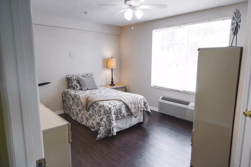 A small bedroom with a single bed covered in a patterned bedspread and pillows. There is a nightstand with a lamp next to the bed, a window with blinds letting in natural light, a wall-mounted air conditioning unit below the window, and a tall dresser on the right side of the room. The floor is dark wood, and the walls are painted light beige.