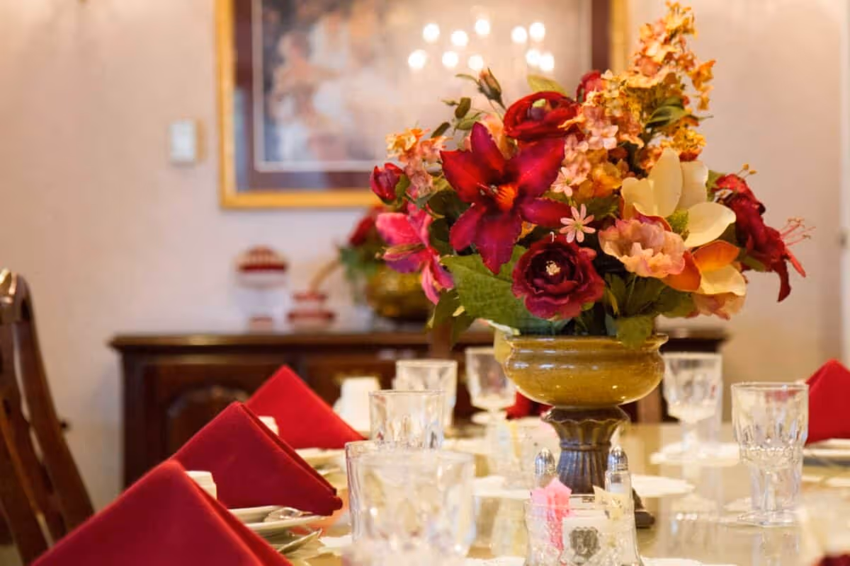 A dining table set with red folded napkins, clear glassware, and a large floral centerpiece featuring red, pink, and yellow flowers in a decorative vase. In the background, there is a wooden sideboard and a framed painting on the wall.