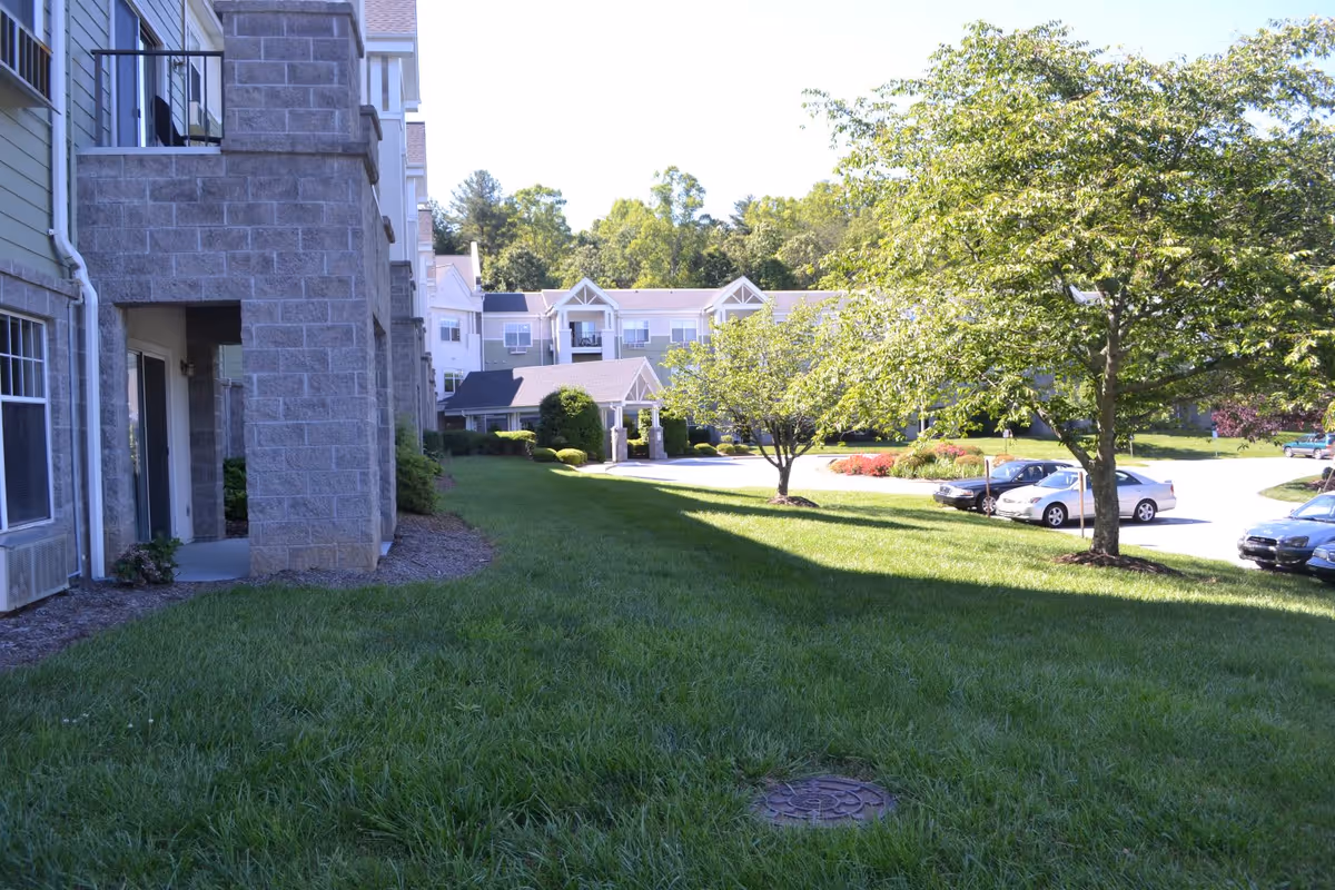 Outdoor view of Trinity View Retirement Community showing a grassy area with trees, parked cars, and a multi-story building with balconies and stone pillars.
