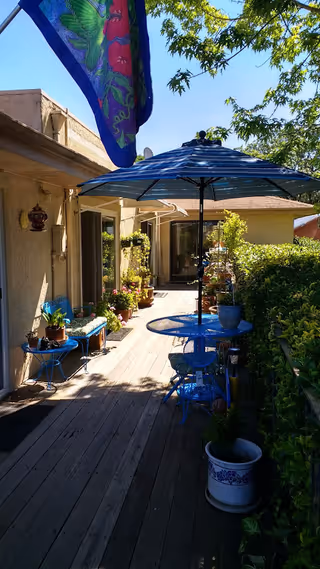 Wooden deck patio beside a house with a blue bistro table and umbrella, potted plants, and seating under trees.