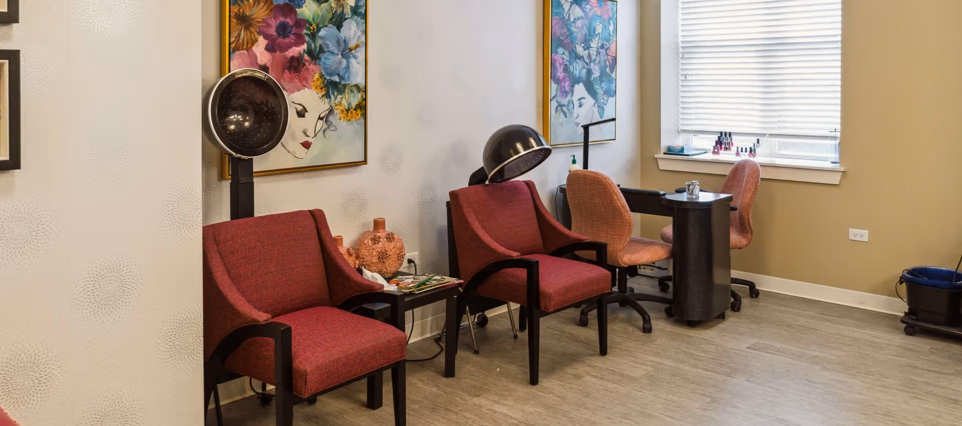 Small salon area with red chairs, hooded hair dryers, a manicure table, and colorful floral artwork.