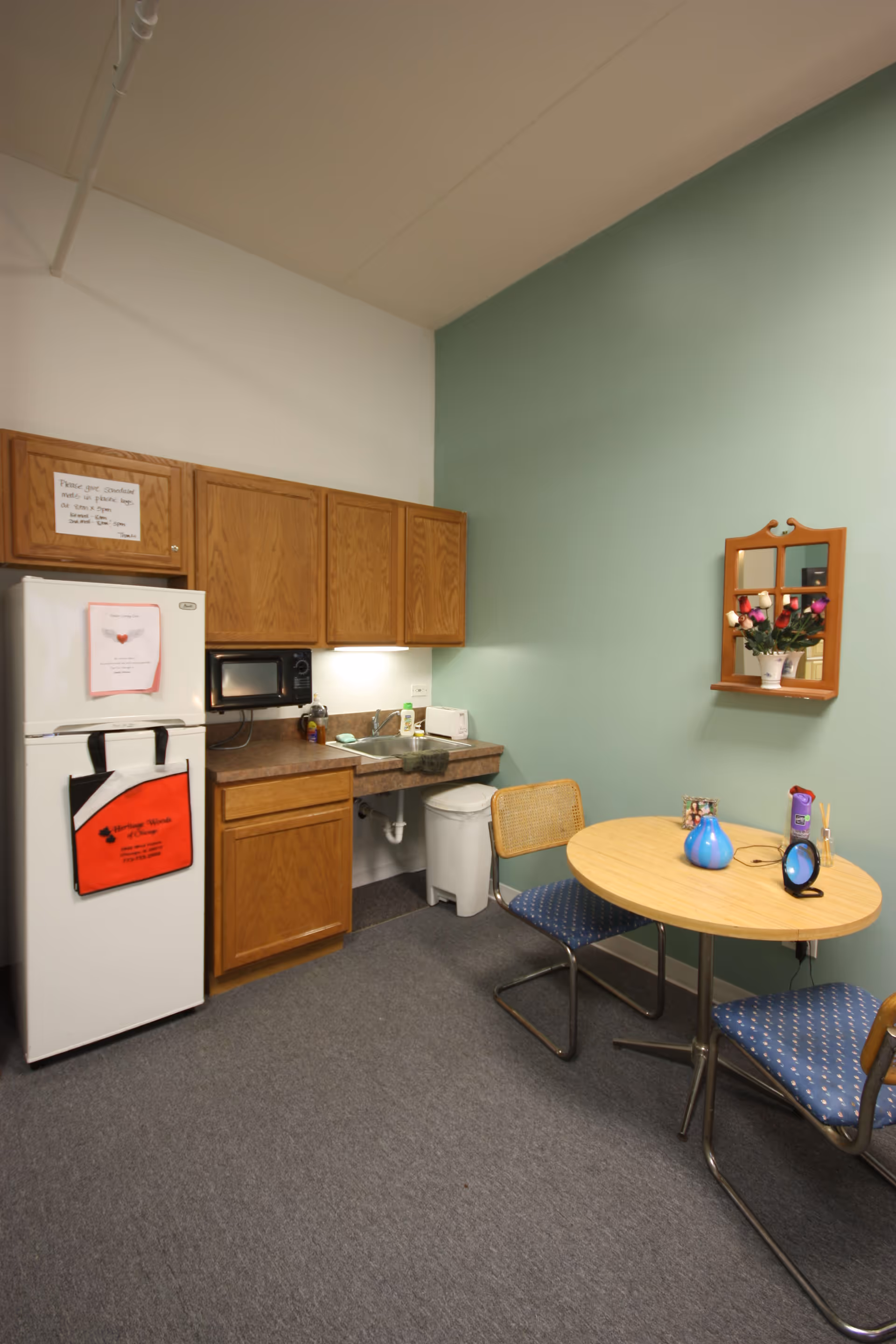 Small kitchenette area with wooden cabinets, a white refrigerator with a red Heritage Woods of Chicago bag hanging on the handle, a microwave, a sink, and a trash can. A round wooden table with two chairs is placed near a green wall with a small wooden mirror shelf holding a vase of colorful artificial flowers.