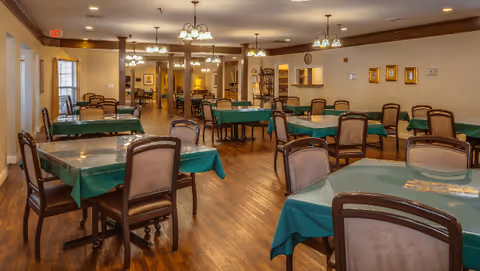 A spacious dining room with multiple tables covered in green tablecloths and surrounded by wooden chairs with cushioned seats. The room has wooden flooring, warm lighting from ceiling fixtures, and framed pictures on the walls. There is a doorway and windows visible in the background.