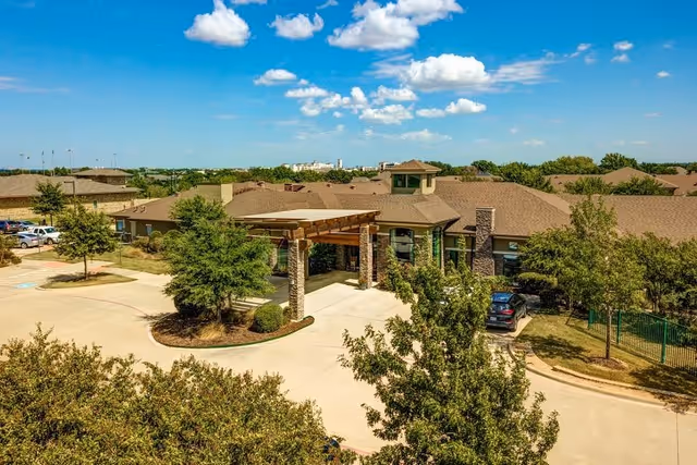Aerial view of the senior living facility entrance with a porte-cochère, surrounding trees, and parking under a blue sky.