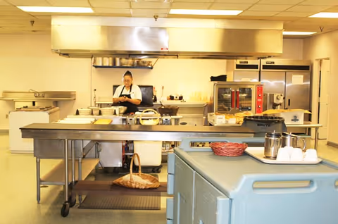 A commercial kitchen with stainless steel counters and appliances. A person wearing a white shirt and black apron is standing behind the counter preparing food. Various kitchen equipment and utensils are visible, including ovens, refrigerators, and a large ventilation hood.