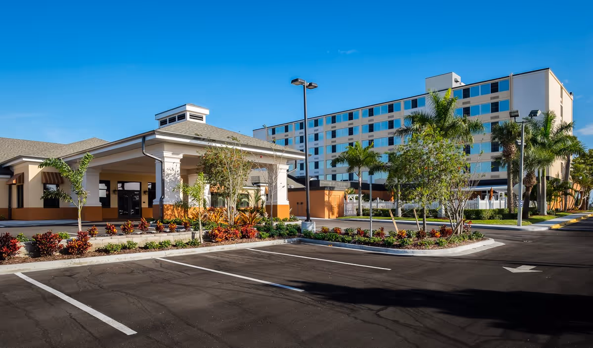 Exterior view of Volante Senior Living of Melbourne facility showing a large multi-story building with many windows, a smaller building with a covered entrance, palm trees, landscaped plants, and an empty parking lot under a clear blue sky.