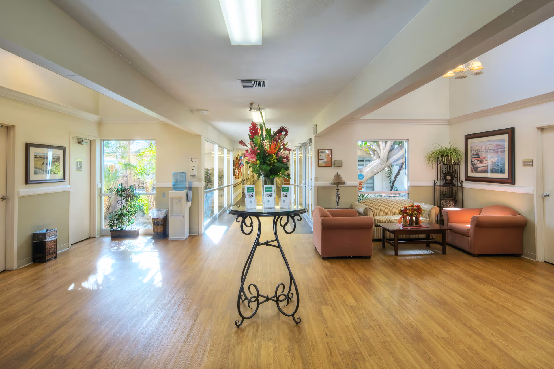 A bright and spacious interior common area with wooden flooring, featuring a round table with a floral arrangement in the center. To the right, there is a seating area with a sofa, two armchairs, a coffee table with flowers, a floor lamp, and framed artwork on the walls. Large windows allow natural light to fill the space, and a water cooler is visible near the entrance on the left side.