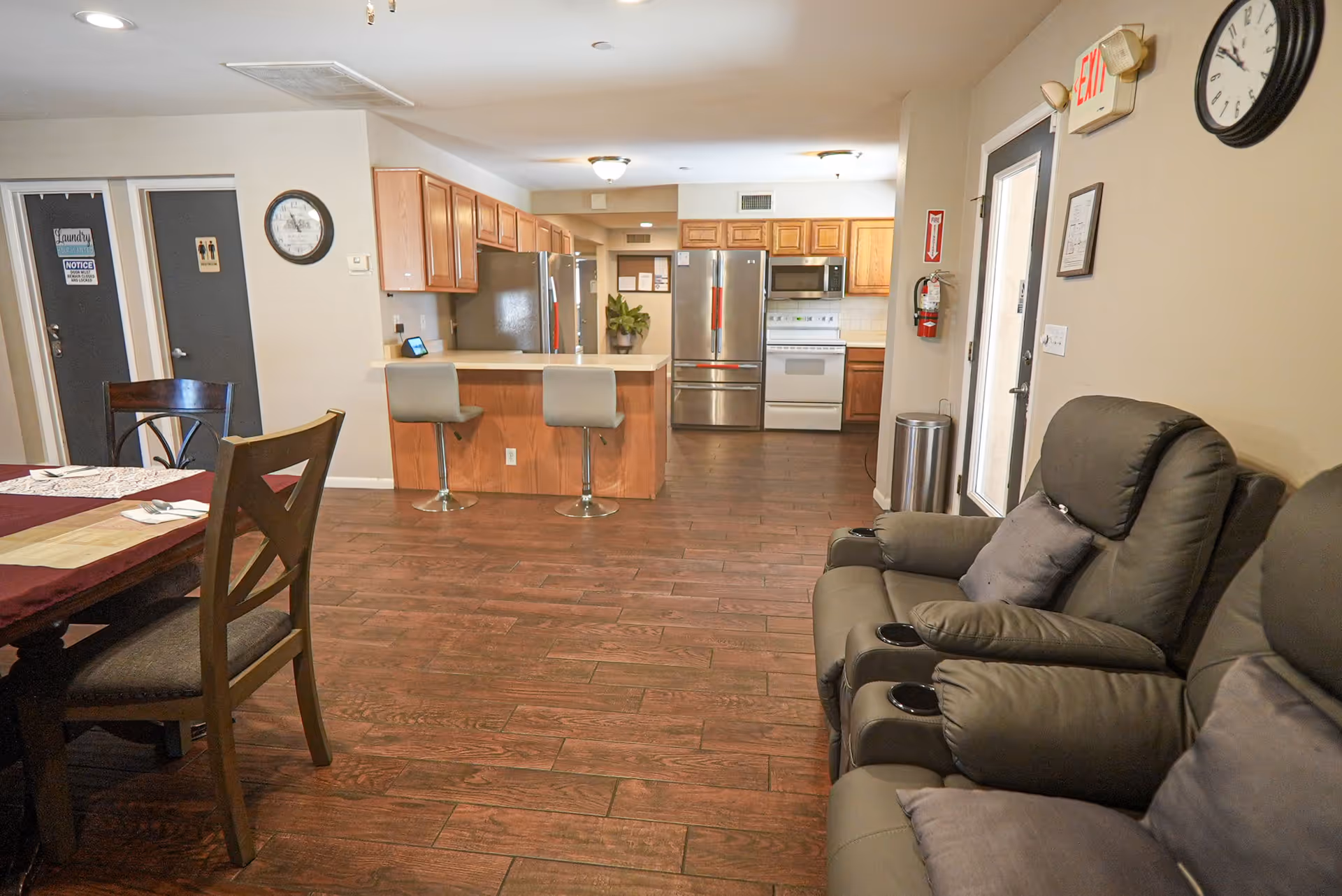 Interior view of a senior living facility showing a kitchen area with wooden cabinets, stainless steel refrigerator, stove, and microwave. In front of the kitchen is a counter with two bar stools. To the left, there is a dining table with chairs, and to the right, two reclining chairs with cushions. The floor is wooden, and there are doors labeled for laundry and restrooms in the background. A clock and an exit sign are visible on the walls.