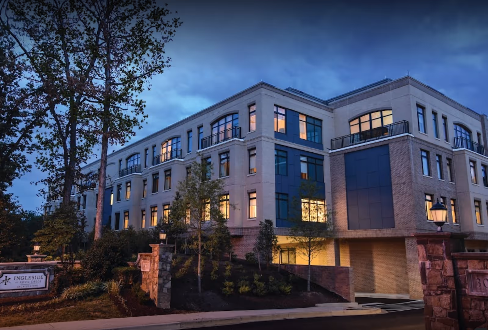 Exterior view of a multi-story senior living facility building named Ingleside at Rock Creek during dusk, with lights glowing from the windows and surrounded by trees and landscaping.