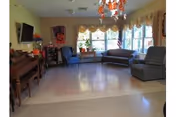 A bright communal living room with sofas, armchairs, a piano, wall-mounted TV, chandelier, and large bay windows with valances.