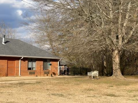 Brick single-story building next to a large yard with a bench and a leafless tree under a partly cloudy sky.