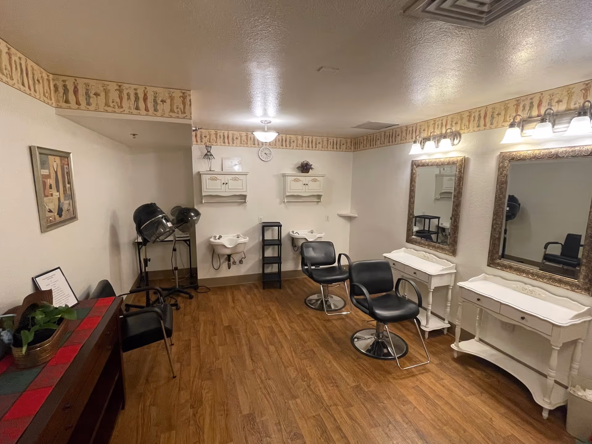Interior of a salon room with two black salon chairs in front of two large ornate mirrors mounted on the wall. Below each mirror is a white vanity table. On the opposite wall, there are two white sinks with shelves above them. The room has wood flooring, a decorative border near the ceiling, and a couple of hair drying machines in the corner.