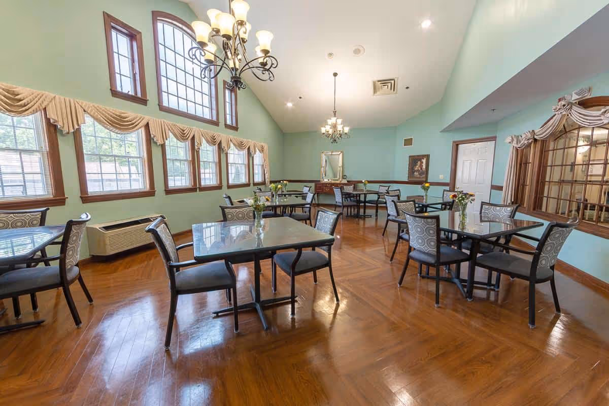 Bright dining room with multiple tables and chairs, large windows with valances, chandeliers, and polished wood floors.