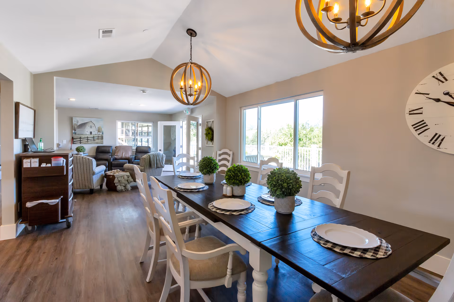 A bright and spacious dining area with a dark wooden table set for six, featuring white chairs and small green potted plants as centerpieces. A large window allows natural light to fill the room. In the background, there is a living area with armchairs, a sofa, and a painting on the wall. The room has wooden flooring and two modern spherical chandeliers hanging from the ceiling.