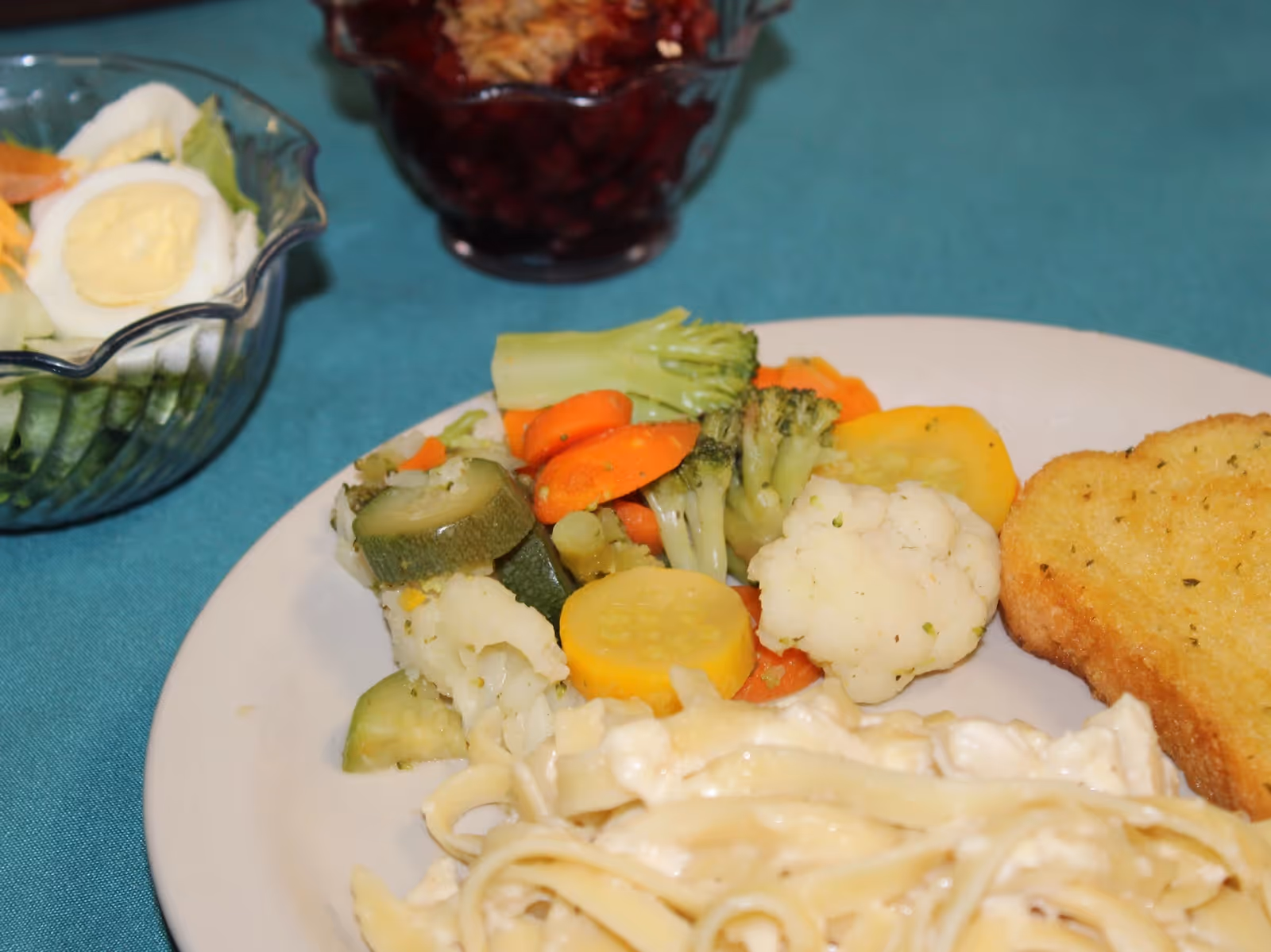 A close-up view of a meal on a white plate featuring cooked mixed vegetables including broccoli, carrots, zucchini, cauliflower, and yellow squash, a slice of garlic bread, and a serving of creamy pasta. In the background, there is a glass bowl with a salad containing sliced cucumbers, lettuce, carrots, and a hard-boiled egg, and another bowl with a red gelatin dessert topped with whipped cream.
