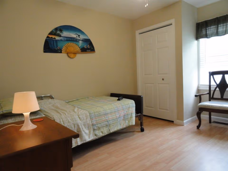 A simple bedroom with a single bed covered in a plaid bedspread, a wooden nightstand with a small lamp, a decorative fan with a beach scene hanging on the beige wall, a white closet door, and a wooden chair near a window with a dark curtain valance.