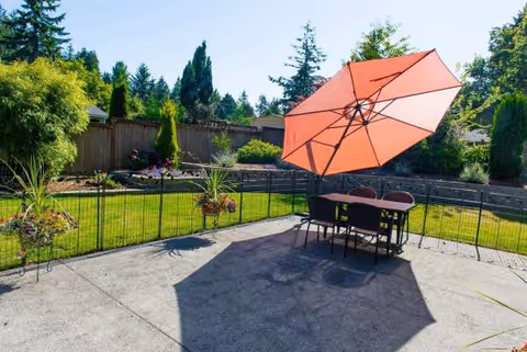 Outdoor patio with table, chairs, and a large orange umbrella in a fenced yard.
