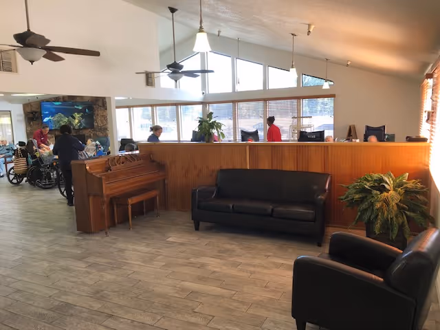 Interior view of a senior living facility common area with a black leather couch and armchair, a wooden piano, several people in the background including one in a wheelchair, large windows letting in natural light, ceiling fans, and plants.